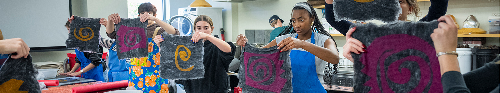 A row of students hold up their handmade felt projects to share with one another during a studio class.