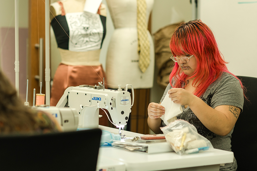 A student sewing at a Juki sewing machine in a studio classroom. Body forms with garments are in the background.