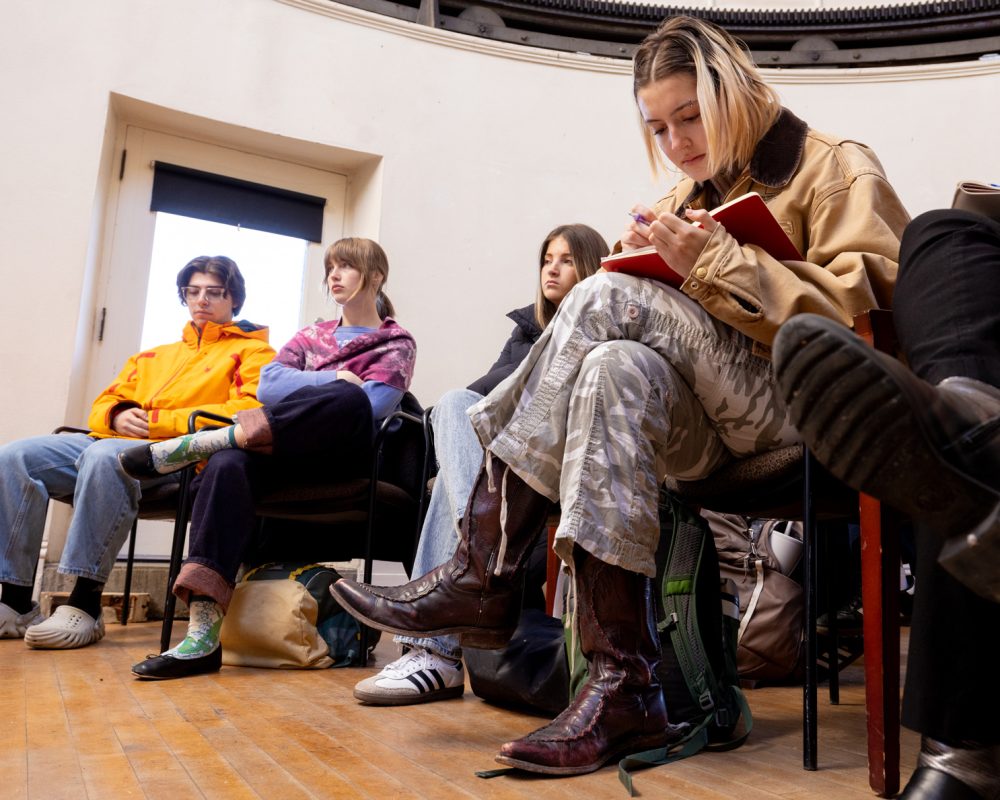 A group of students sit in chairs in a white room. One of them is sitting leaning towards the camera, jotting down notes in a red notebook.