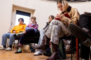 A group of students sit in chairs in a white room. One of them is sitting leaning towards the camera, jotting down notes in a red notebook.