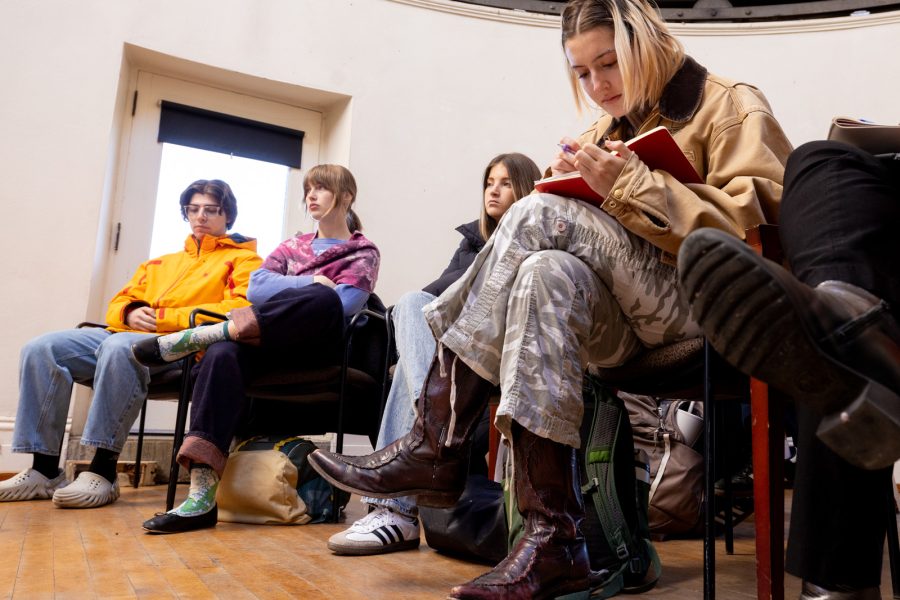 A group of students sit in chairs in a white room. One of them is sitting leaning towards the camera, jotting down notes in a red notebook.