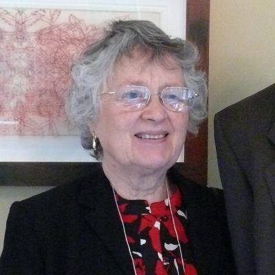 A woman smiling, with short, gray hair, wearing glasses, a red patterned blouse and a black blazer.
