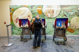 A woman wearing jeans, a backpack and a pony tail, faces a voting machine (with her back to the camera), head down, as she casts a ballot, with a mosaic mural in the background.