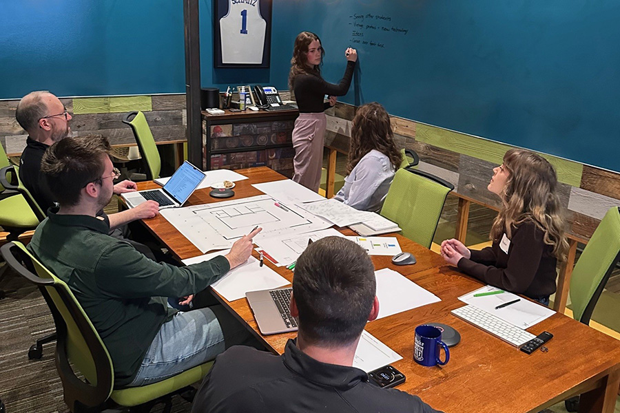 Team collaborating in a conference room as a woman writes on a whiteboard while others review plans and laptops.