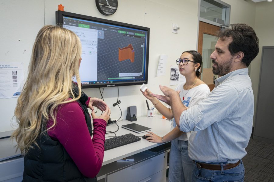 3 students discuss a 3D model on screen while preparing a part for the lab's 3D printer.