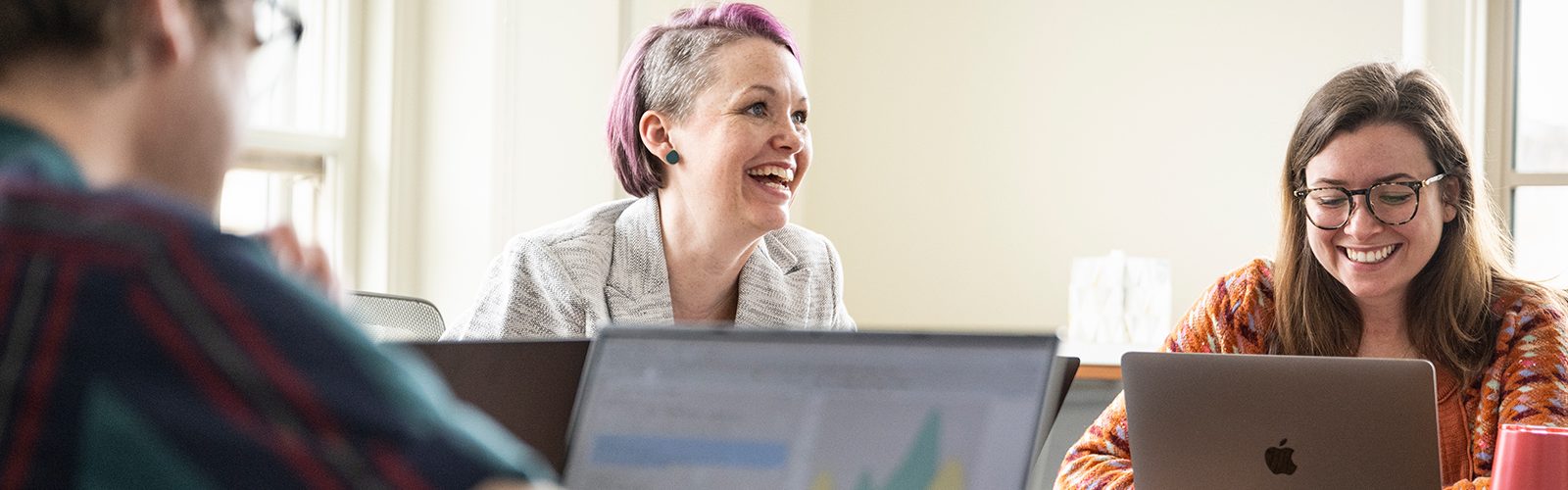 A faculty member smiles while sitting at a conference table with graduate students and their laptops.