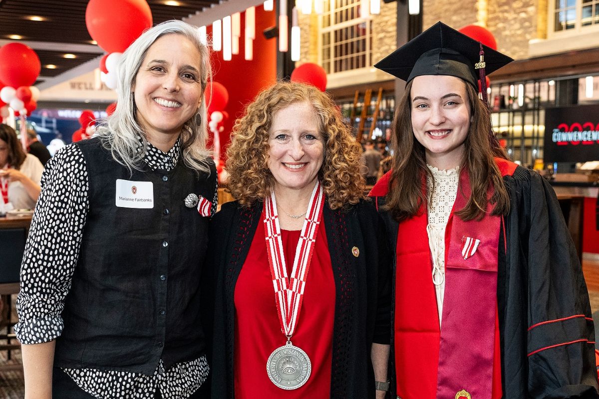 Three people posing indoors at a graduation event, one wearing a cap and gown with a red stole, another with a silver medal around her neck.