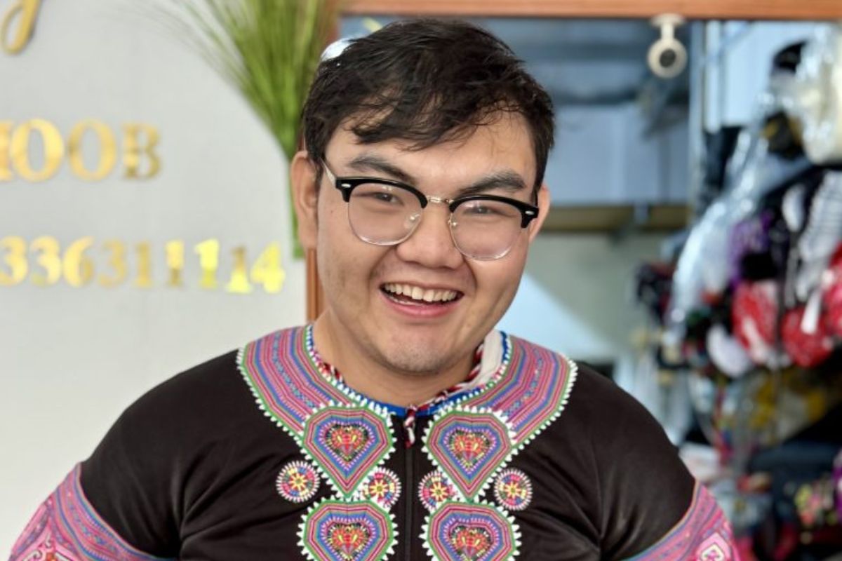 A young man with black hair and glasses smiles and wears a vibrant shirt with magenta stitching.