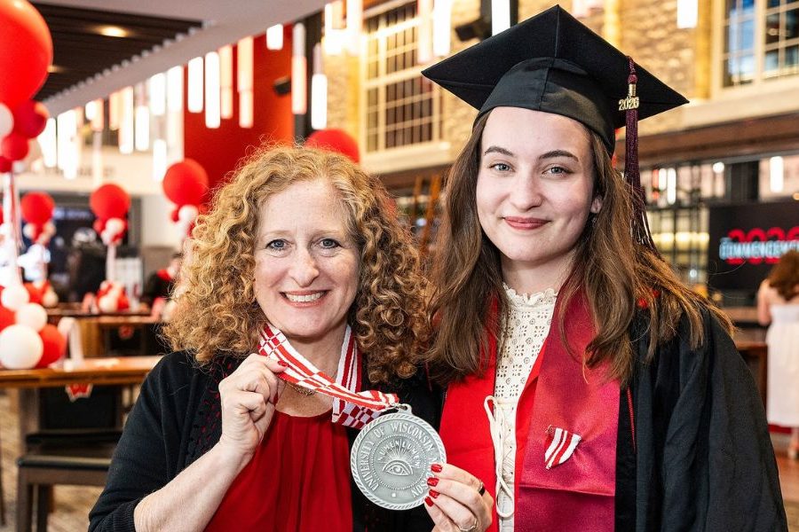 Graduate in cap and gown stands beside a woman holding a silver medal at a festive indoor celebration with red and white balloons.