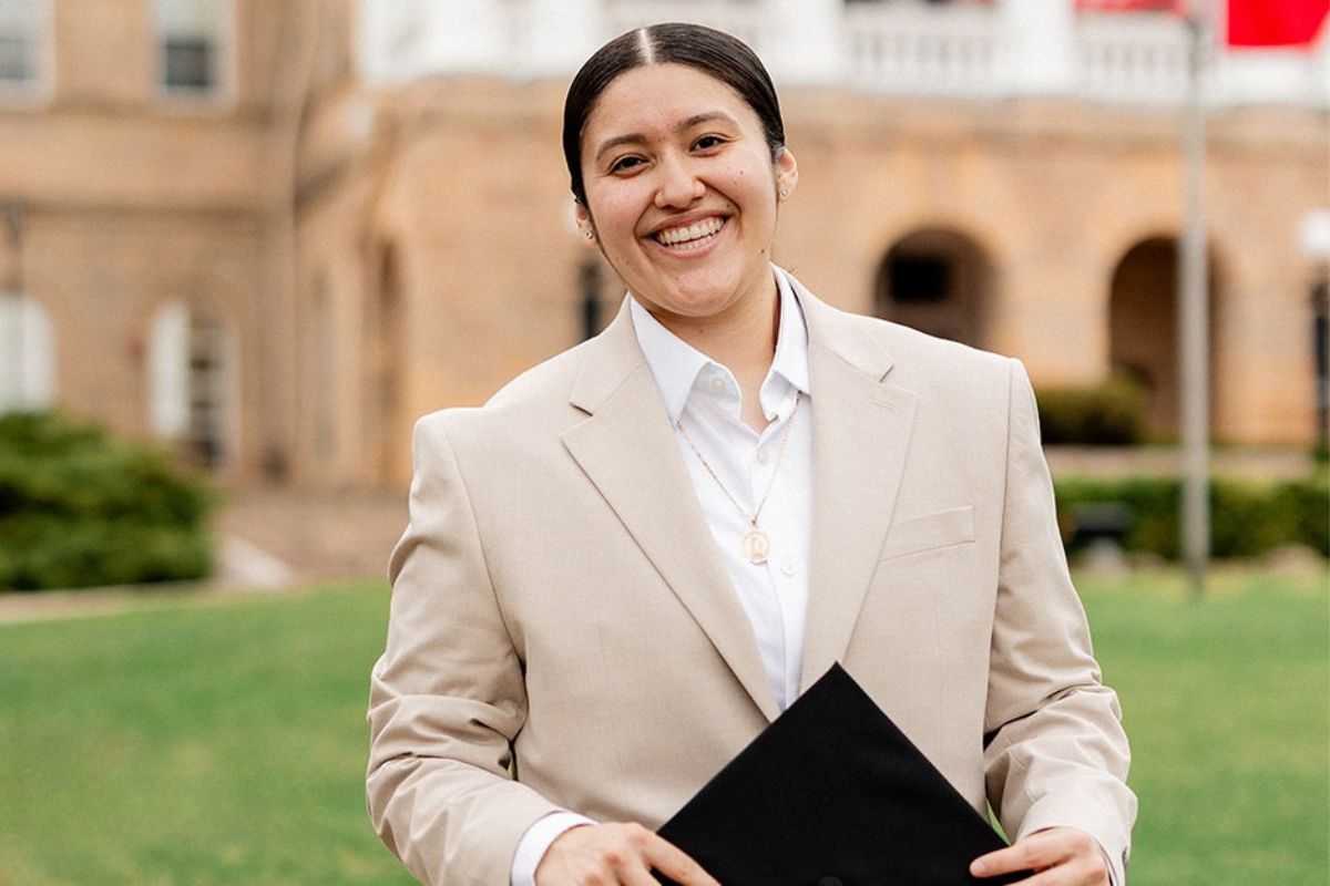 A young woman with pulled back black hair wearing a tan suit, holds a black graduation cap.