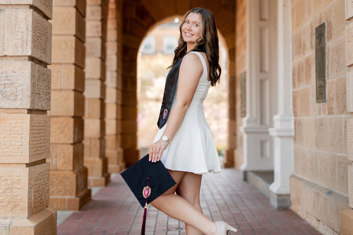 A young woman poses in a white dress and black graduation stole while holding a black graduation cap with the UW–Madison crest.