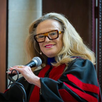 An older woman smiling, with blonde hair framing her face, wearing large, rectangular tortoiseshell glasses, holds a microphone with her left hand and looks at the camera. She is wearing a black ceremonial robe with red accents.