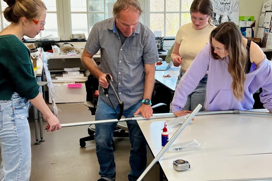 Design instructor demonstrating frame-fabrication techniques to three attentive students gathered around a studio worktable