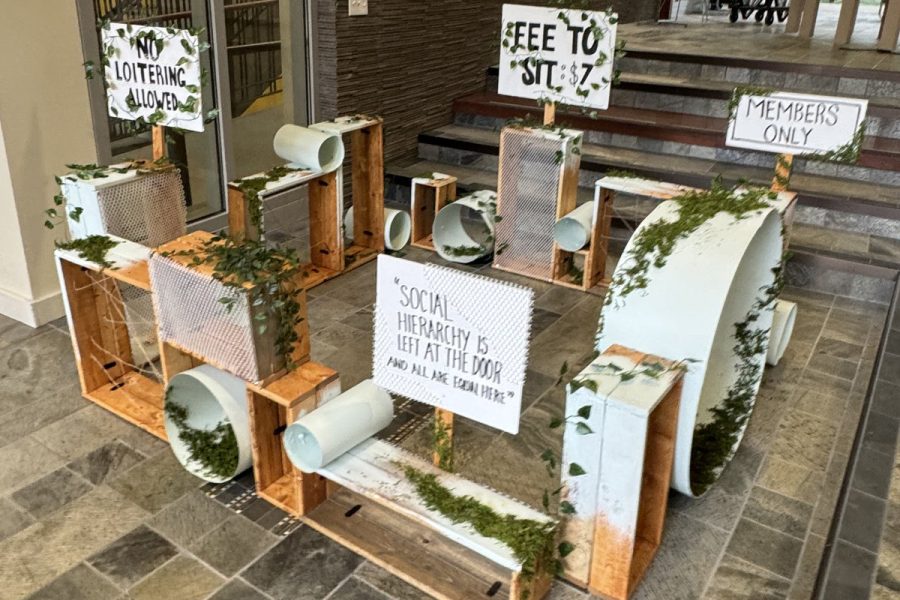 Student-built wooden crate installation in a building stairwell featuring greenery and hand-lettered signs that critique social hierarchy and exclusion