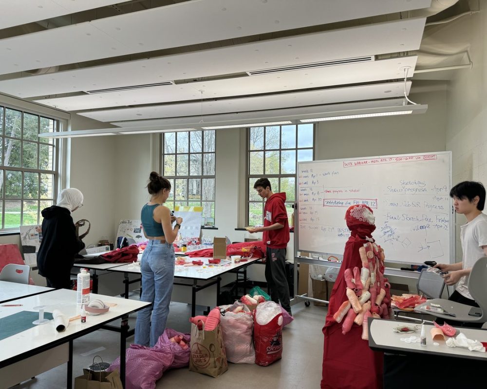 Students in a design classroom working together on a large red fabric sculpture surrounded by tools, sketches, and bags of materials