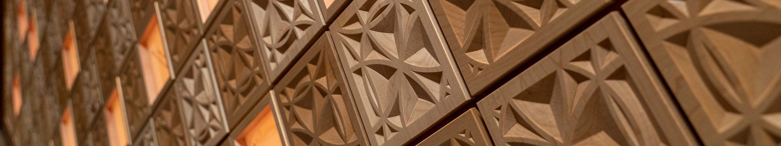 Close up of the wall in Ember Hall, composed of ornately carved, wooden square diamond shapes. Amongst the tiles are red, glowing diamonds displaying donor names