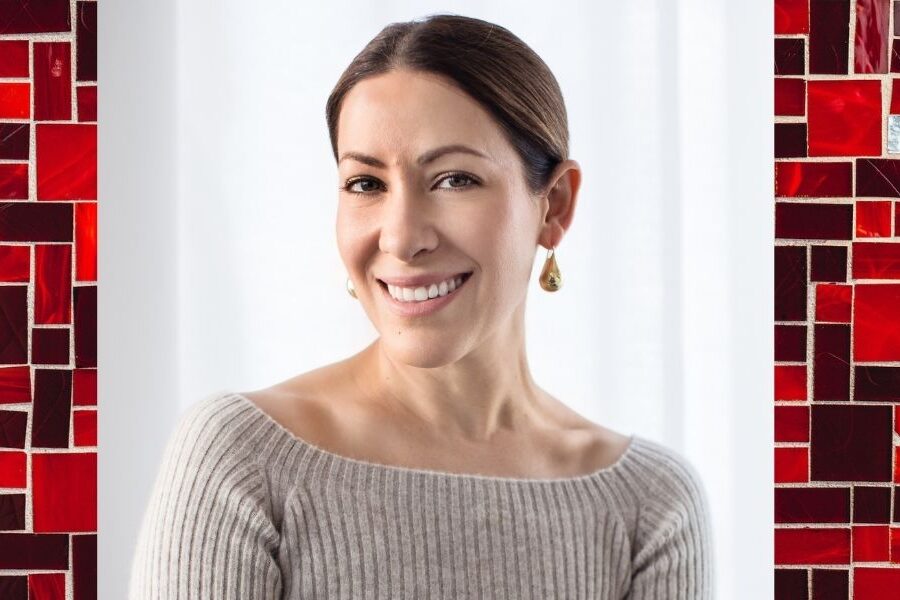 A woman wearing a light sweater with brown hair pulled back into a bun smiles for a photo. Photo is set on a backdrop of red, black and white tiles.