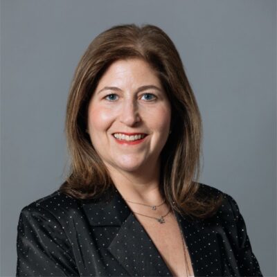 A white woman smiling, with brown, straight hair, wearing a black blazer.