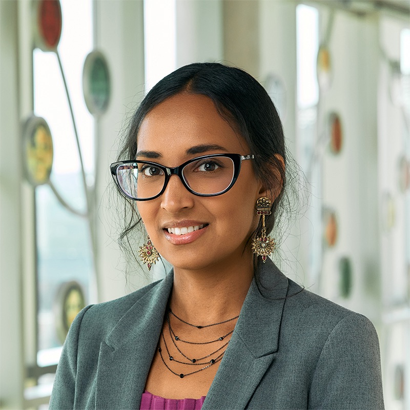 An Indian-American woman smiling, with black hair clipped back, wearing a deep pink dress, grey blazer, black necklace, and bronze earrings.
