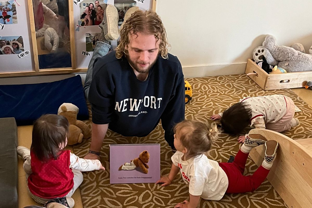 An adult surrounded by three toddlers doing yoga poses on the floor of a carpeted classroom. A teddy bear diagram showing the pose is in the middle.
