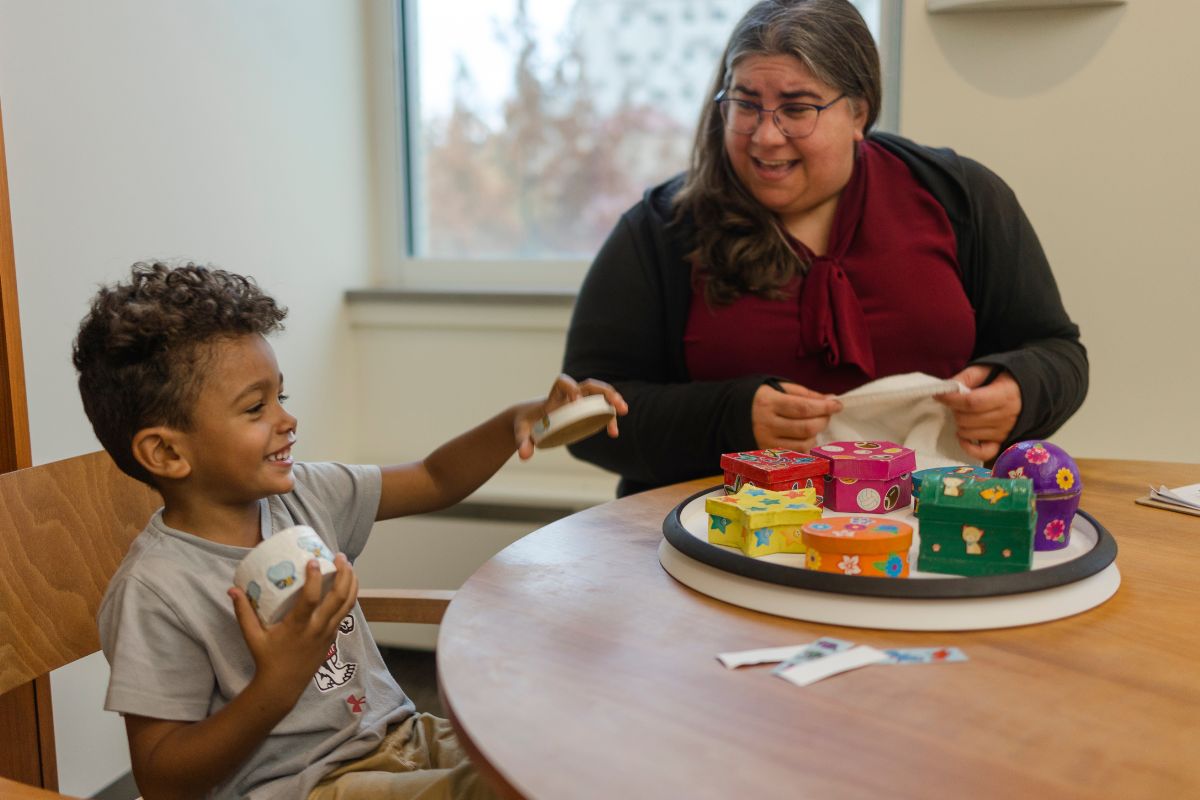 An adult smiles at a smiling child holding a small white box they opened. The two are sitting at a table with a turntable with assorted colored boxes on it.
