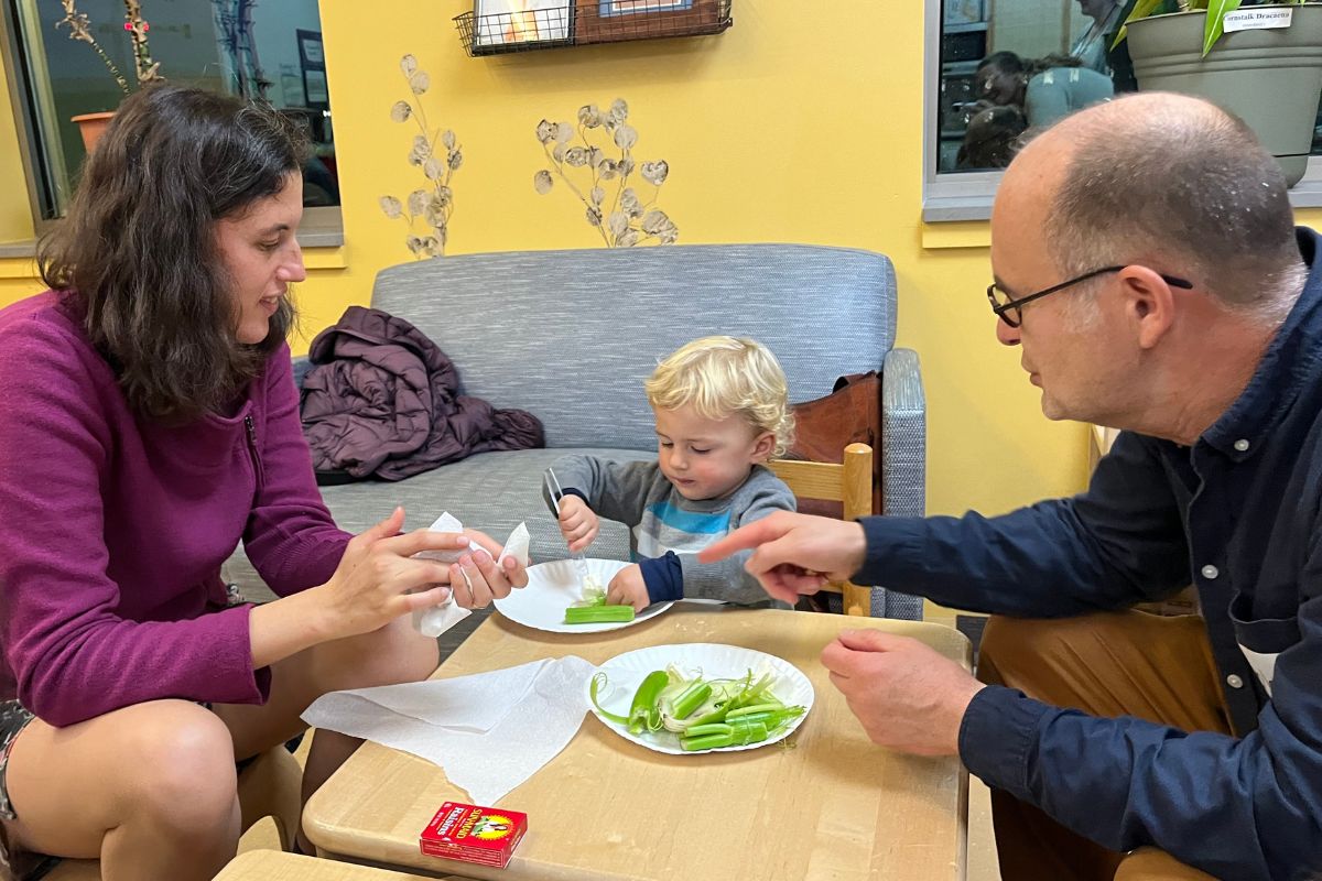 Two adults sit at a small table with a child and instruct them to spread cream cheese onto celery.