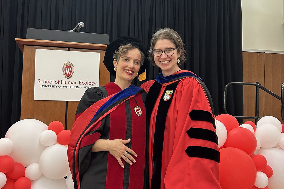 Two people in red, black and dark blue commencement gowns stand together and smile in front of a stage. On the stage is a large red and white balloon sculpture and a wooden lecturn with a microphone.