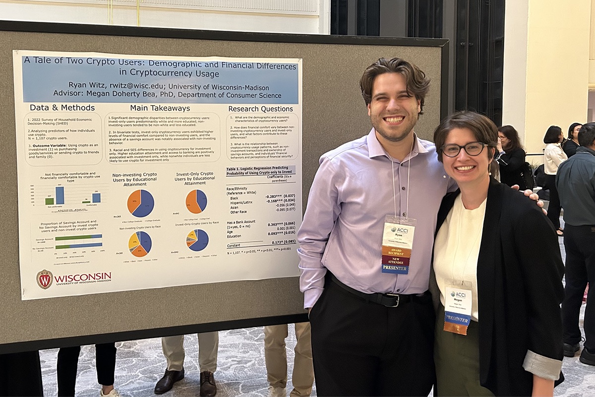 Two people wearing professional clothing pose together and smile in front of a large board displaying a research poster titled "A Tale of Two Crypto Users: Demographic and Financial Differences in Cryptocurrency Usage."
