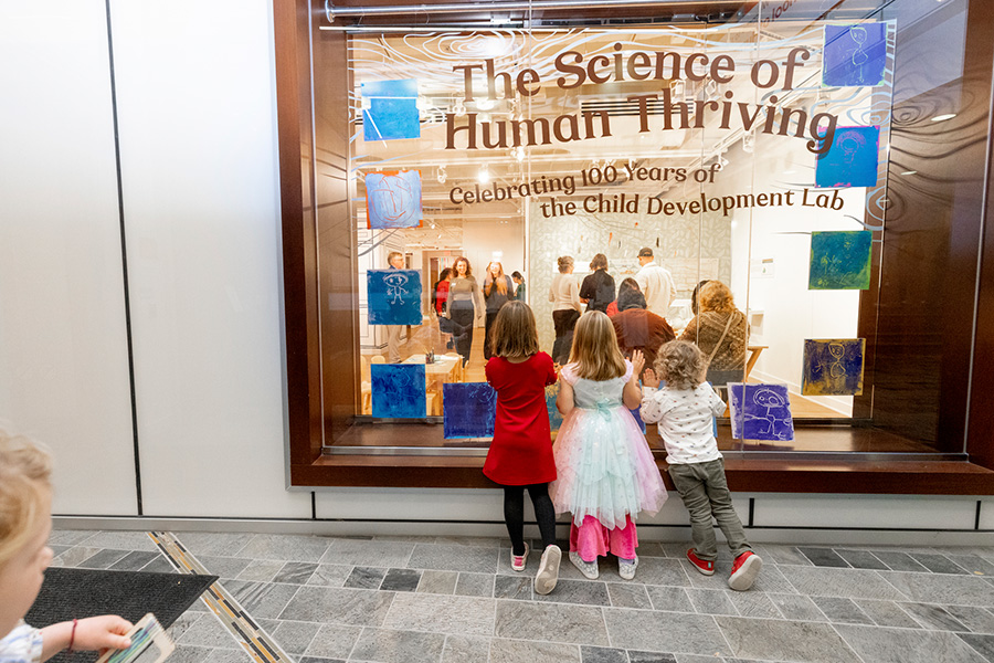 Three children peer through a gallery window that says “The Science of Human Thriving Celebrating 100 years of the Child Development Lab".