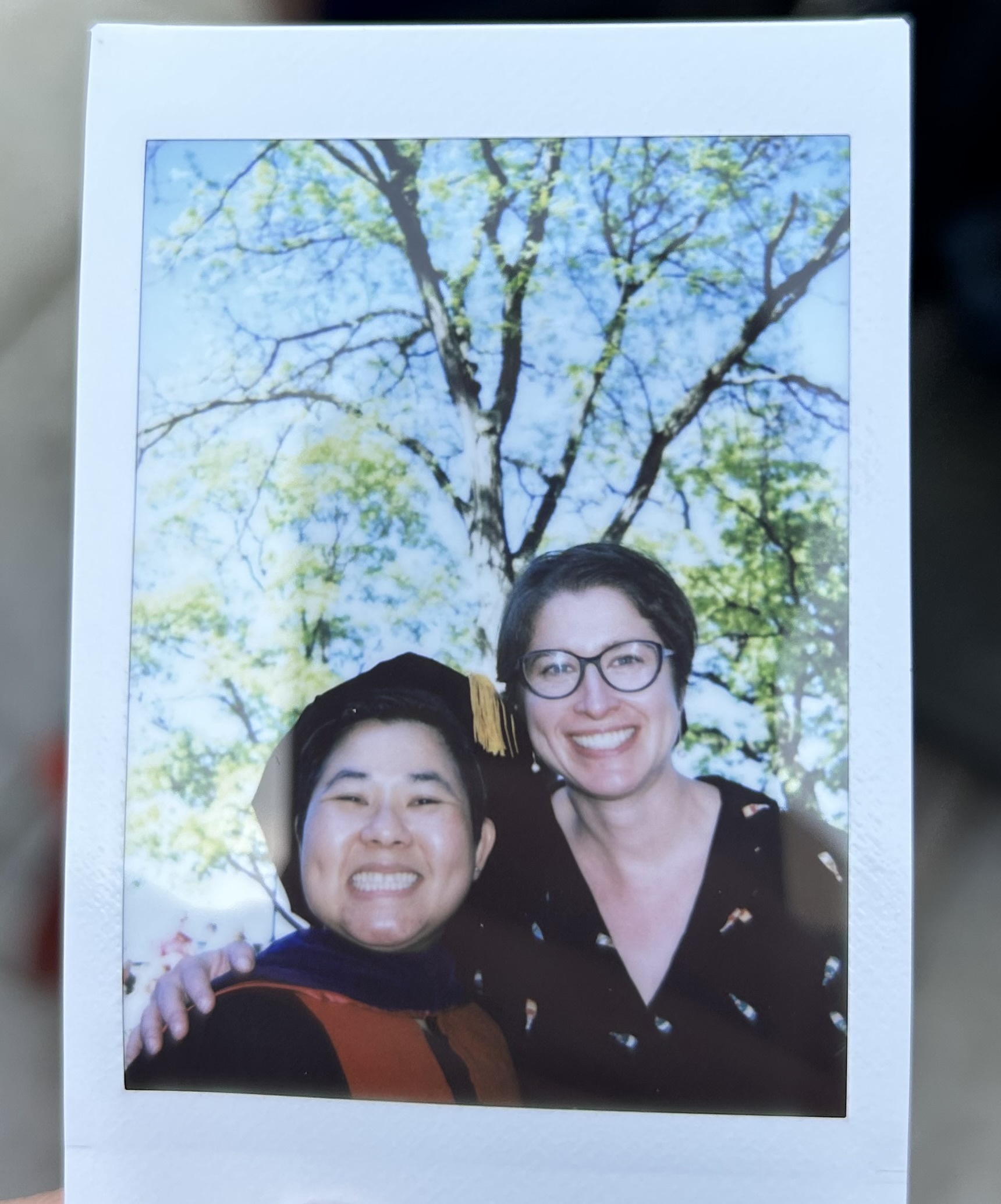 A photo showing a Polaroid picture of two people posing together and smiling under a blue sky and a tree with green leaves. One of the people is wearing a commencement gown and hat.